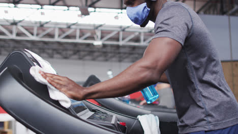 fit african american man wearing face mask and headphones cleaning treadmill machine with disinfecta