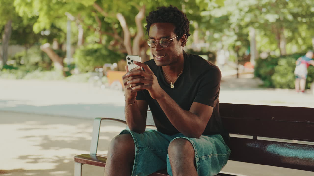 Giovane uomo afroamericano sorridente mentre usa lo smartphone in un parco.