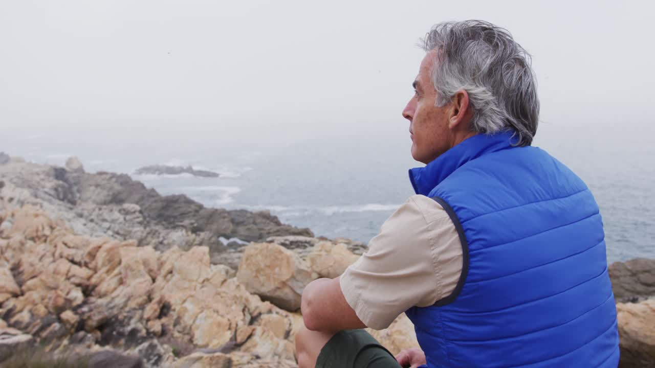 Senderista mayor sentado en las rocas en las montañas observando la costa.