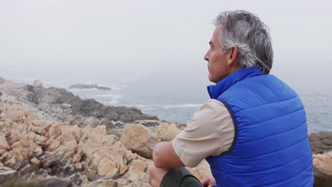 Senior-hiker-man-sitting-on-the-rocks-in-the-mountains-and-watching-the-sea-shore.