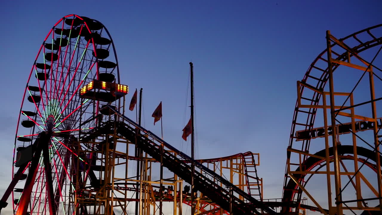 Drapeaux flottant au vent près d'une grande roue et de montagnes russes dans un parc d'attractions au crépuscule.