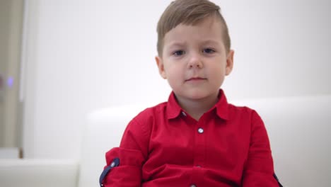 Cheerful-little-boy-sitting-on-the-sofa-and-playing-with-a-smartphone-gadget.-Looking-at-the-camera,-smiling-and-showing-his-cool-boots-with-lights.-Shot-in-4k