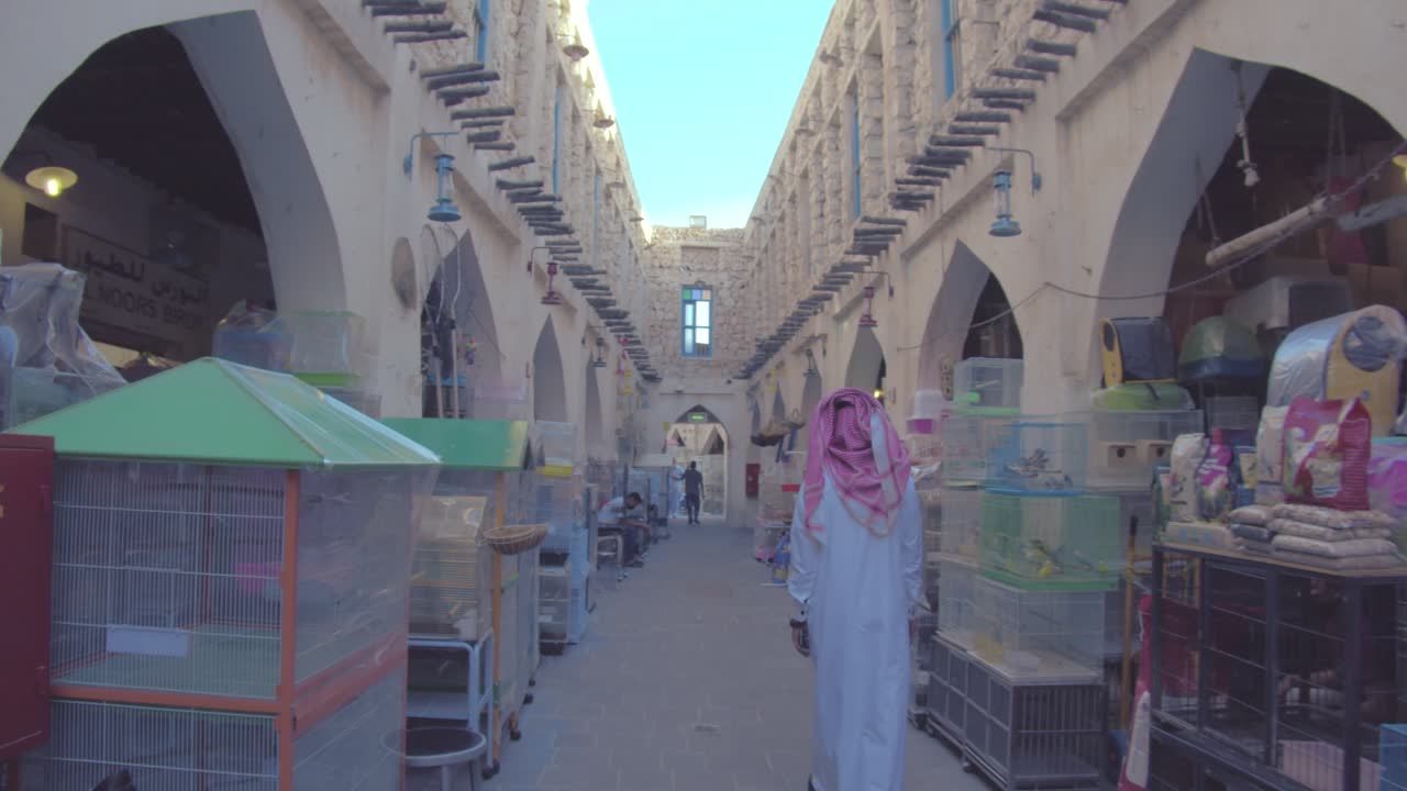 Back View Of An Arab Man In White Thawb And Agal Passing By A Pet Shop In Souq Waqif, Doha, Qatar. handheld shot