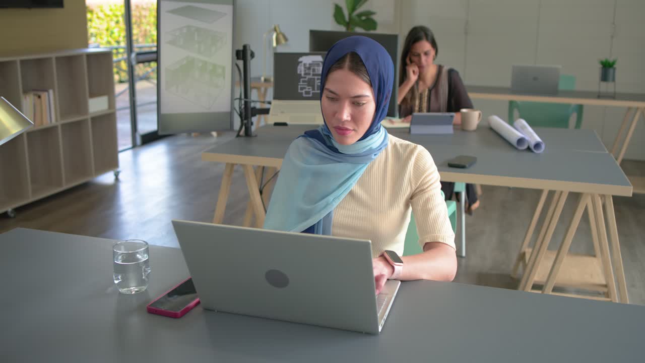 Rekan kerja asal India mengetik di laptop sambil memeriksa tablet di kantor, berhenti sejenak, berbalik, dan mendiskusikan proyek