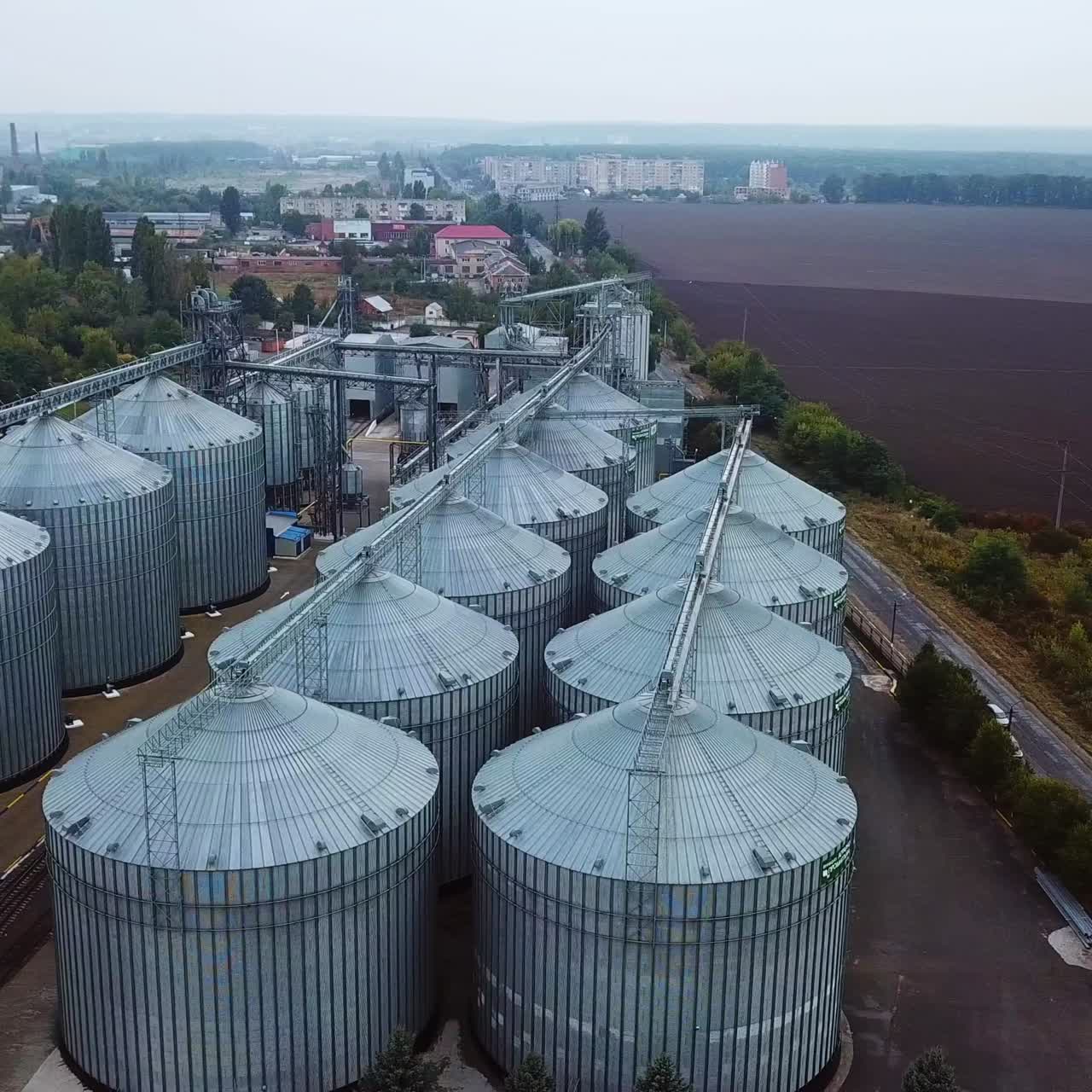 Enormous metal tanks standing in straight rows. Railways dividing the plant territory. Cityscape and farmlands at backdrop