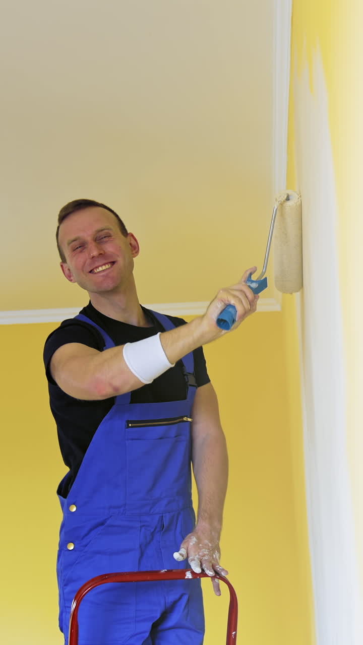 Man in work overalls paints the wall. Workman in the uniforms painting the walls with paint roller. Vertical video