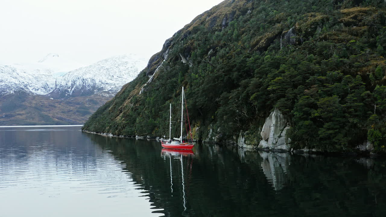 Red sailboat anchored in narrow fjord along Beagle Channel surrounded by green forest cliffs, angled approach