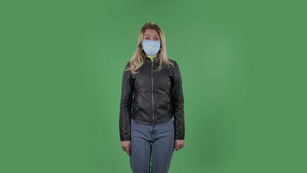 Portrait of beautiful young woman in medical protective face mask looking at camera worries and scared. Blonde with loose hair in a black jacket and jeans on a green screen in the studio. Health Protection Corona Virus Concept