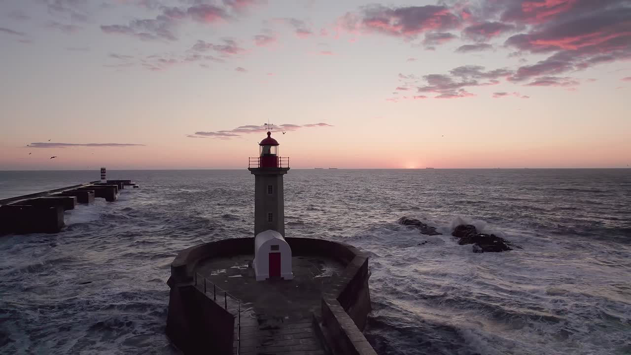 Drone orbits Farol da Barra lighthouse standing on rocky breakwater at Rio Douro estuary mouth during sunset, Atlantic waves surrounding fortified structure with horizon clouds glowing amber