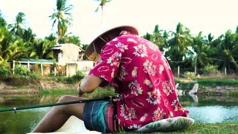 a fisherman sitting on the ground is wearing a hat, a motley t-shirt, and shorts