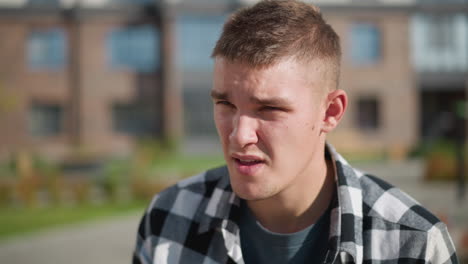 white man wearing checkered shirt closes eyes after taking medication while sitting outdoors on bright sunny day, with blurred background showing office building and greenery