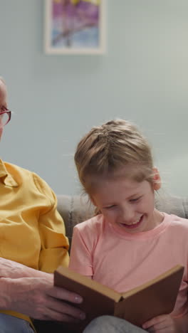 blonde grandma and granddaughter laugh at funny moment in book sitting together on couch in living room slow motion. family generations spend time at home