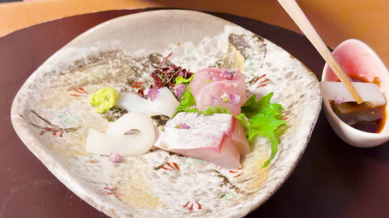 陶器の皿に盛られた醤油に、木の箸で生のイカを浸しているクローズアップショット。