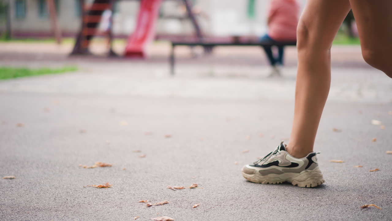 Spor ayakkabılı parkta yürüyen bacaklar, bank ve oyun alanı yakınında kentsel bir yolda; rahat bir yaşam tarzı ve hafif bir fitness rutinini yansıtan rahat bir adım, sıcak yaz ışığı ve sığ alan derinliği, yakın çekim odak.