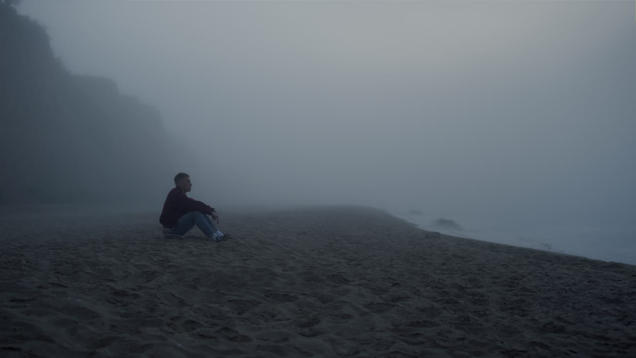 Sad man looking sea landscape in fog. Serious guy sitting on sandy beach