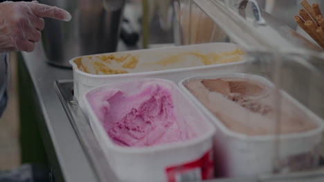 primer plano de varios recipientes de helado con diferentes sabores, servidos por una persona con guantes