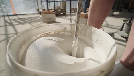 a construction worker mixes mortar with a power drill in a bucket on a scaffolding-covered building site, showcasing the construction process