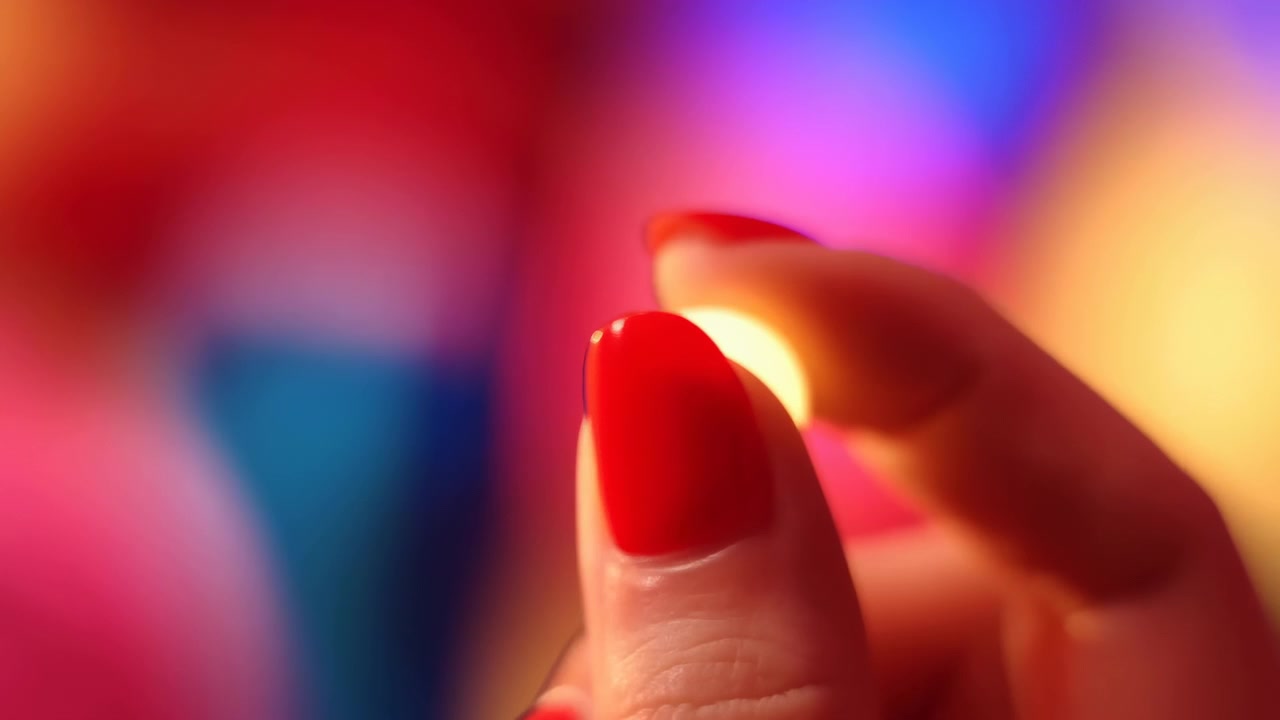Primo piano di una mano con smalto rosso che tiene un oggetto luminoso su uno sfondo vibrante e sfocato, in un'ambientazione di bellezza commerciale misteriosa e intrigante, vista dall'alto.