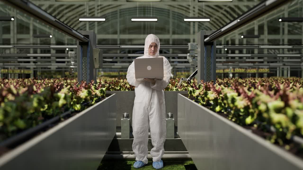 cuerpo lleno de hombre asiático investigador usando una computadora portátil mientras estaba de pie en el invernadero con agricultores robóticos inteligentes