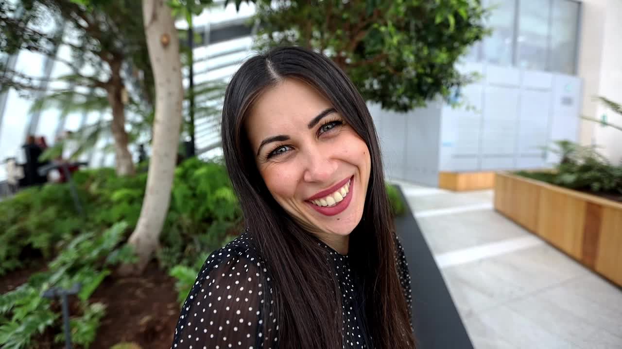 cámara lenta de una mujer morena feliz con el pelo largo sonriendo frente a la cámara en un edificio con jardín