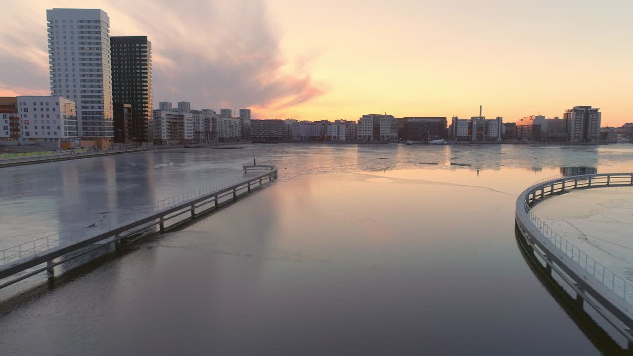 스웨덴 스홀름의 호수 에 있는 현대 아파트 건물의 드론 촬영. 물과 얼음 위를 날아다니는 공중 촬영, 배경에 현대 주거 지역, 일몰