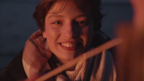 camera focuses on a teenage girl smiling sitting around bonfire with her friends