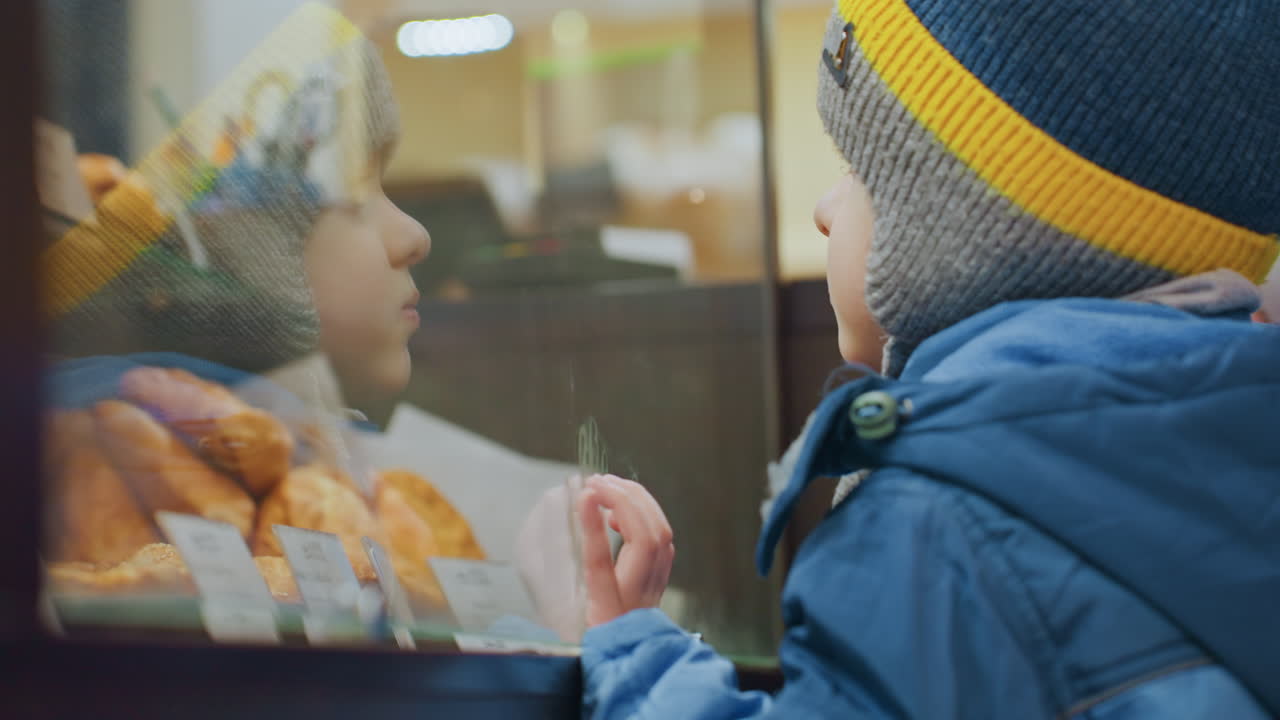 Bambino ammira dolci. Bambino con cappotto blu guarda con calma la selezione di pasticcini attraverso la vetrina della panetteria. Giovane ragazzo con cappotto brillante osserva pacificamente biscotti e torte attraverso la vetrina del negozio.