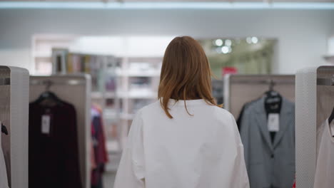 vista trasera de una mujer joven explorando el pasillo de una tienda minorista con estantes de ropa bien dispuestos, mostrando varios estilos y colores en un entorno tranquilo y bien iluminado