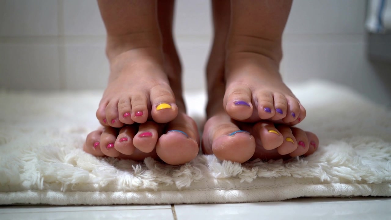 Saliendo del baño, dos pares de pies se asientan en una alfombra color crema, mostrando las uñas pintadas.