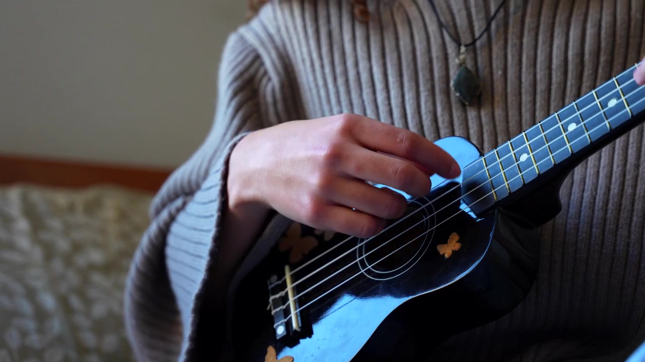 Primo piano delle mani di una giovane donna che strimpella una piccola chitarra acustica in una stanza accogliente