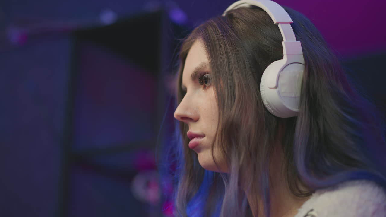 close up side view young girl with long eyelashes wearing white headphones deeply focused on screen with blurred lights in background under soft lighting