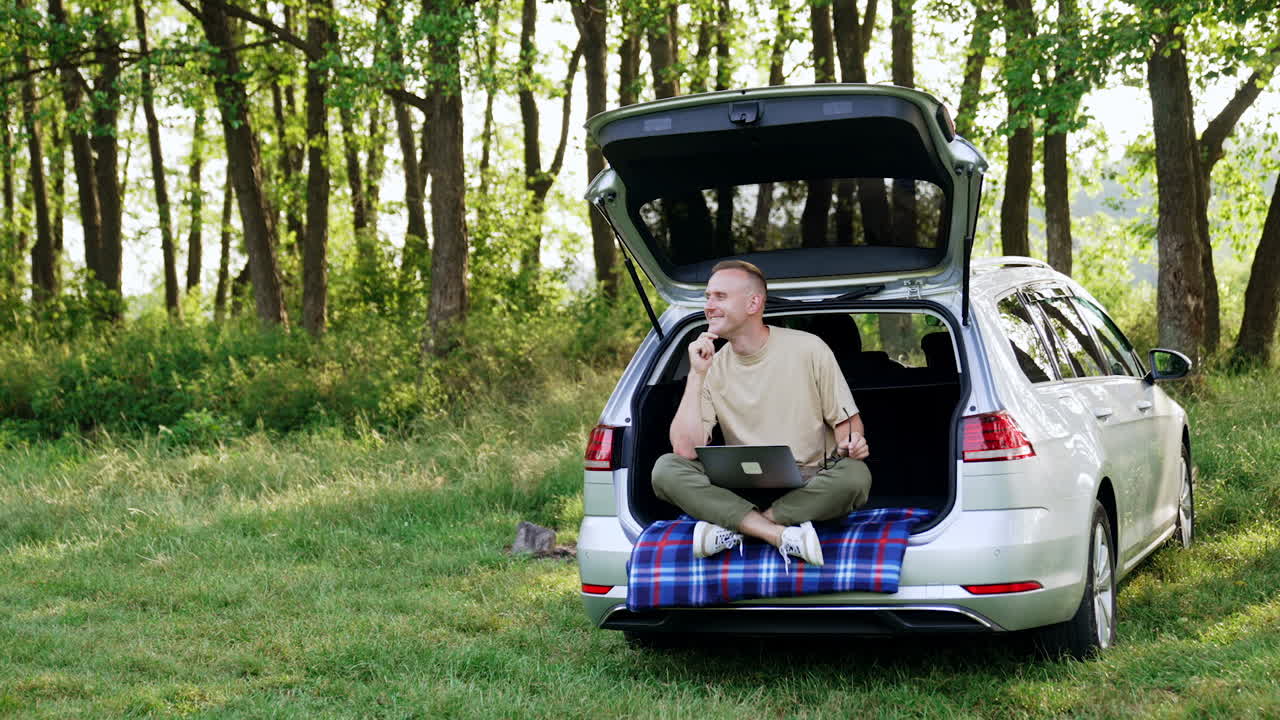 Thoughtful Caucasian man works in nature on his laptop. Happy man sits in car trunk looking pensively around, thinking about ideas.