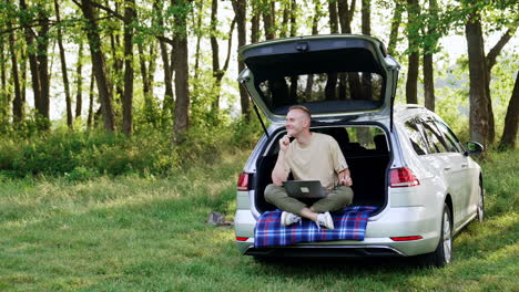 thoughtful caucasian man works in nature on his laptop. happy man sits in car trunk looking pensively around, thinking about ideas.