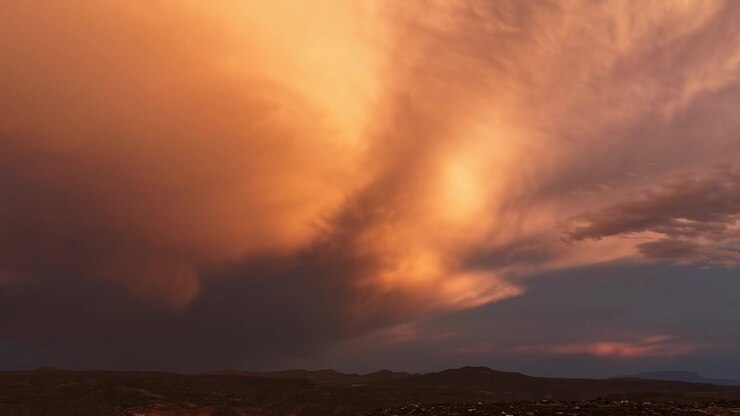 A drone timelapse of a dark monsoon storm clearing over a rugged Southwest canyon at sunset. Vibrant evening colors glow against the heavy clouds, illuminating distant rain shafts across the terrain.
