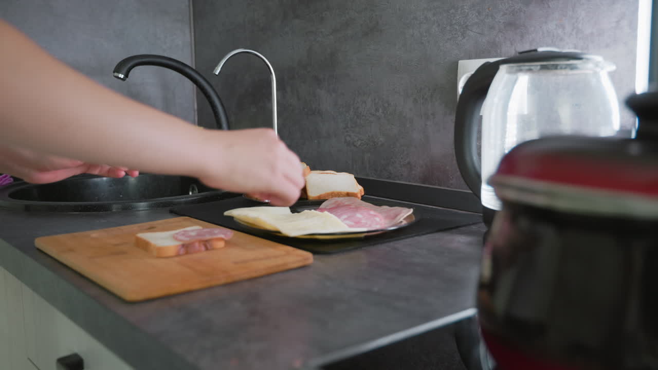 Hands preparing sandwiches with slices of bread, cheese, and deli meat on kitchen counter next to cutting board and electric kettle, highlighting casual breakfast or lunch meal preparation in home kitchen setting