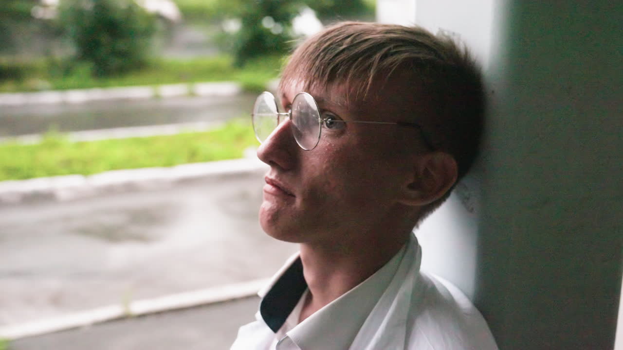 Young biologist in white coat leans head against wall during outdoor break, eating pastry with thoughtful expression in calm urban setting surrounded by greenery and wet pavement