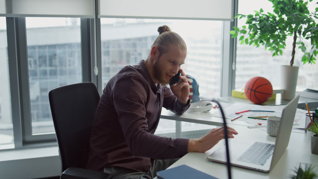Nærbillede af inspireret designer, der taler i smartphone på et moderne kontor. Mand bruger bærbar computer.
