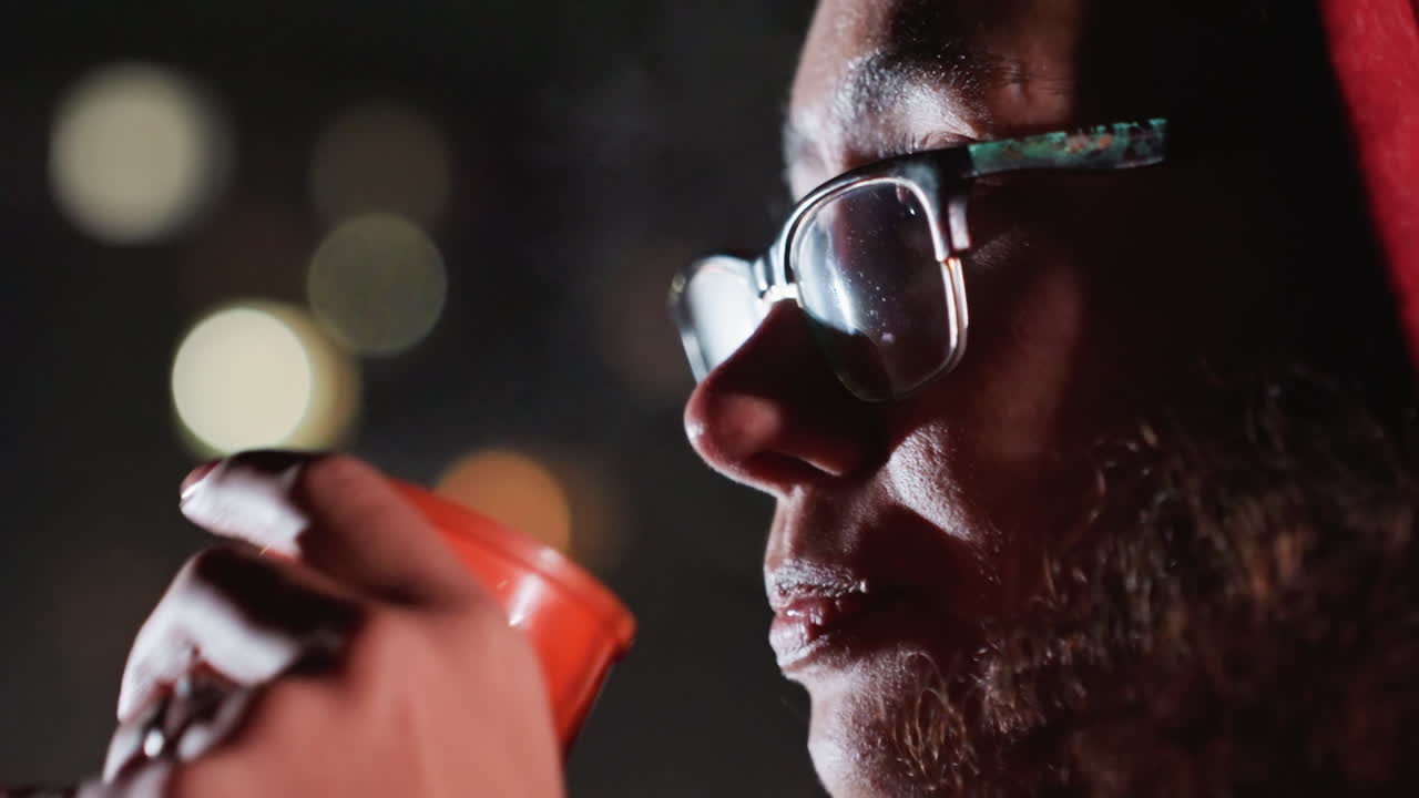 Homme barbu sirotant une boisson chaude en regardant les étoiles tard dans la nuit, astronome amateur se réchauffant les mains, lunettes embuées, bokeh des lumières de la ville, gros plan de profil transmettant une humeur contemplative et tranquille