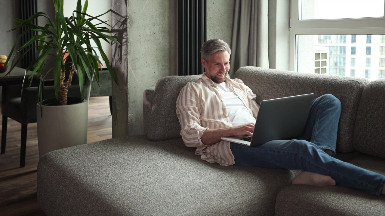 Happy Man Working on Laptop while Relaxing on Sofa at Home