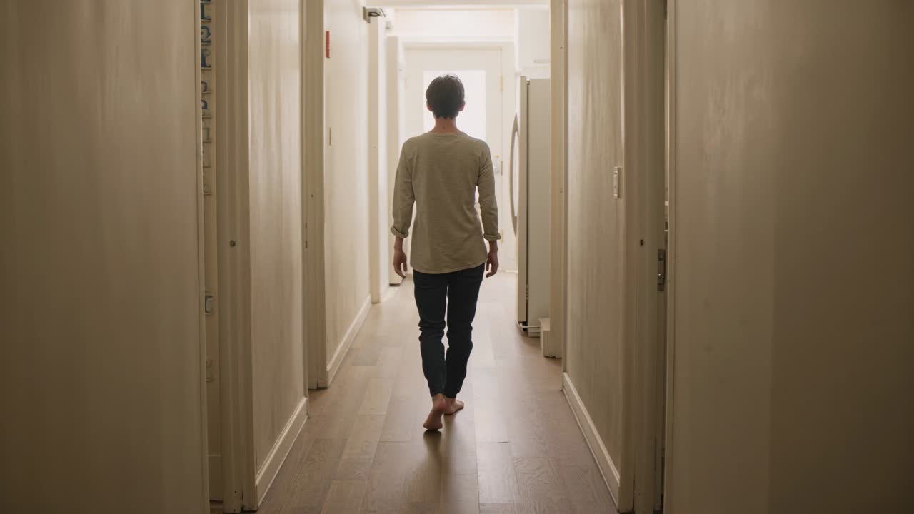 Hombre descalzo caminando con un top claro y pantalones oscuros por el pasillo de casa, pasando la nevera hacia una puerta luminosa.