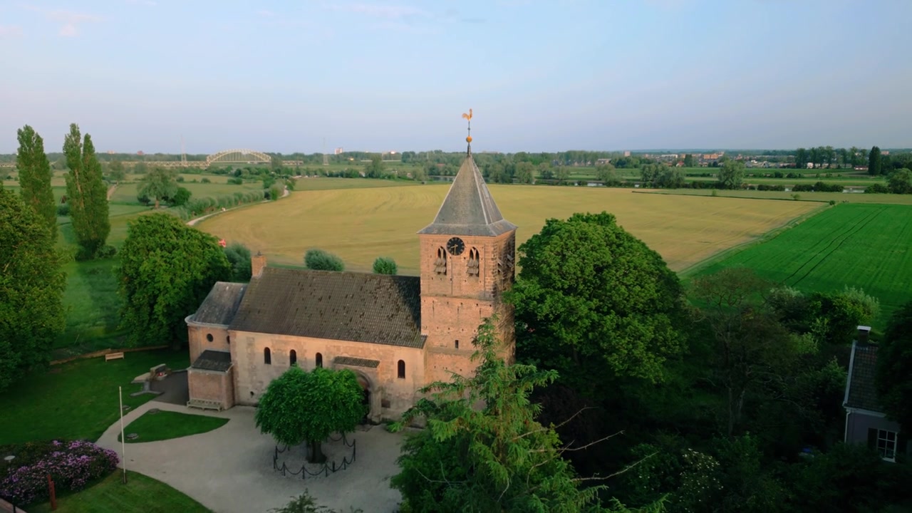 Nahaufnahme aus der Luft des historischen Turms der Alten Kirche von Oosterbeek in den Niederlanden, beleuchtet vom Licht der goldenen Stunde. Zeigt aufwendige architektonische Details, Zifferblätter und eine markante Wetterfahne.