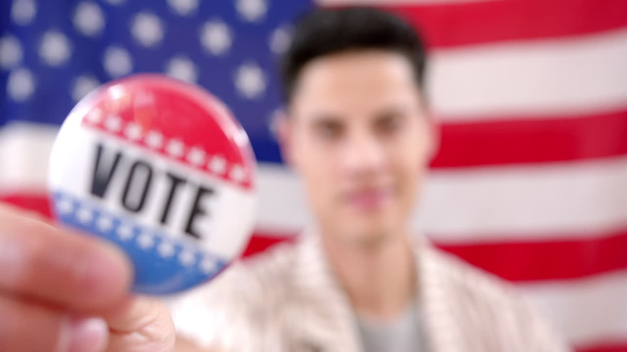Portrait of biracial man in front of american flag holding badge with vote text, slow motion