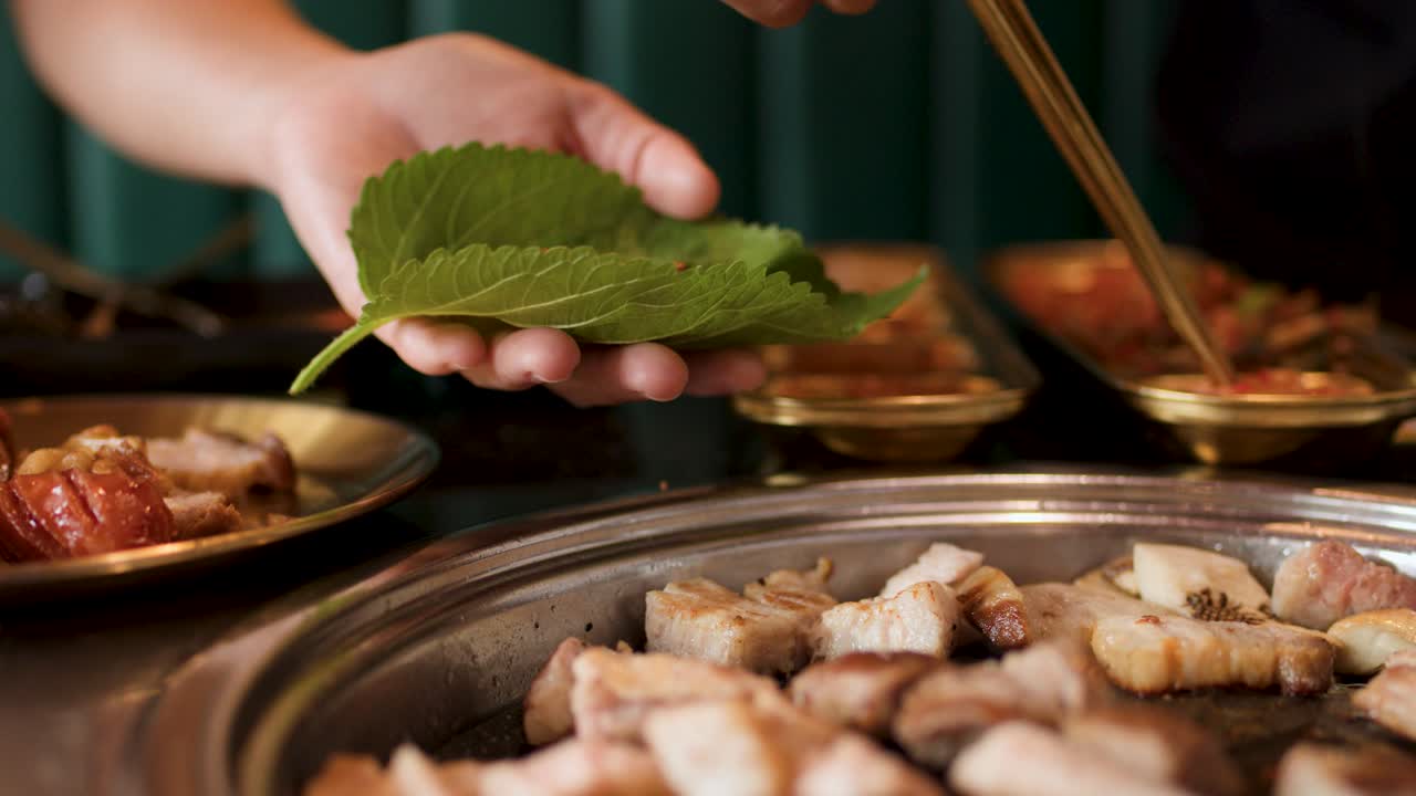 Hand wraps grilled pork and kimchi in perilla leaf using chopsticks at Korean BBQ