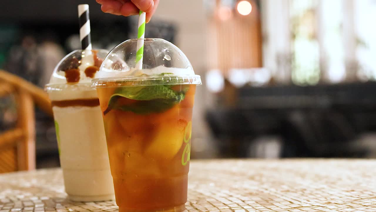 Two iced drinks on a café table are stirred by hand. Bright, natural lighting enhances the refreshing atmosphere