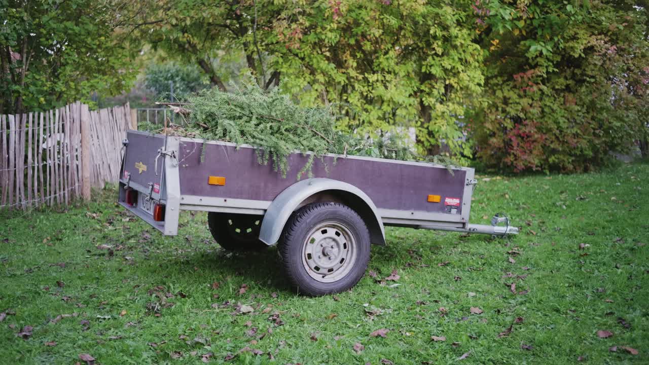 A traditional German garden with a trailer in the background, filled with tree and shrub cuttings, highlighting seasonal yard work and natural greenery