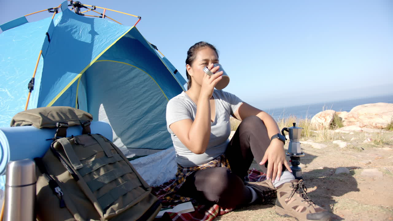 Camping by tent, woman drinking coffee and relaxing after mountain hike