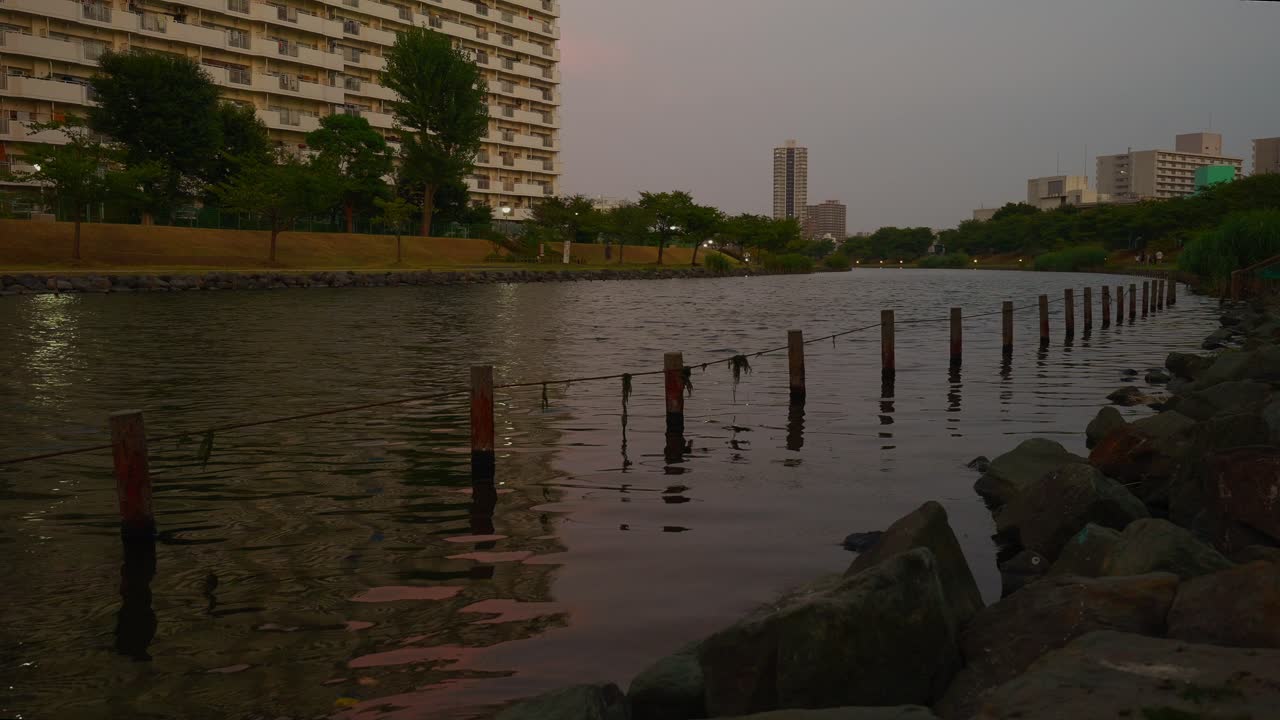 A wide shot of a calm river with a rocky bank in the foreground and city buildings in the distance