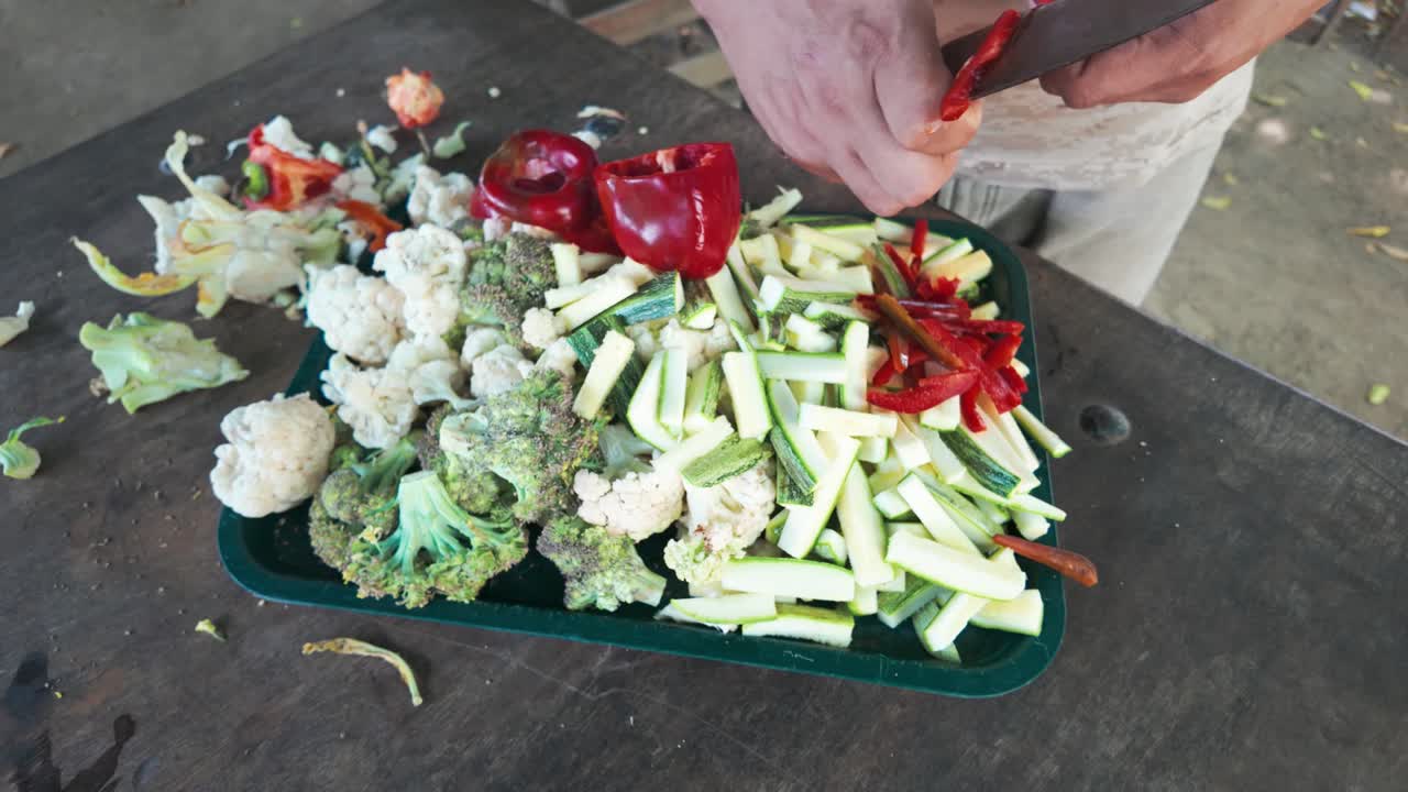 Hands cut red peppers and other vegetables using large knife on a tray, with cauliflower and zucchini