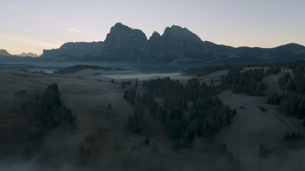 Cinematic aerial view of frosty Alpe di Siusi meadows and scattered mountain huts at dawn, mist drifting across rolling hills beneath the dramatic Sassolungo peaks in the Dolomites, Italian Alps