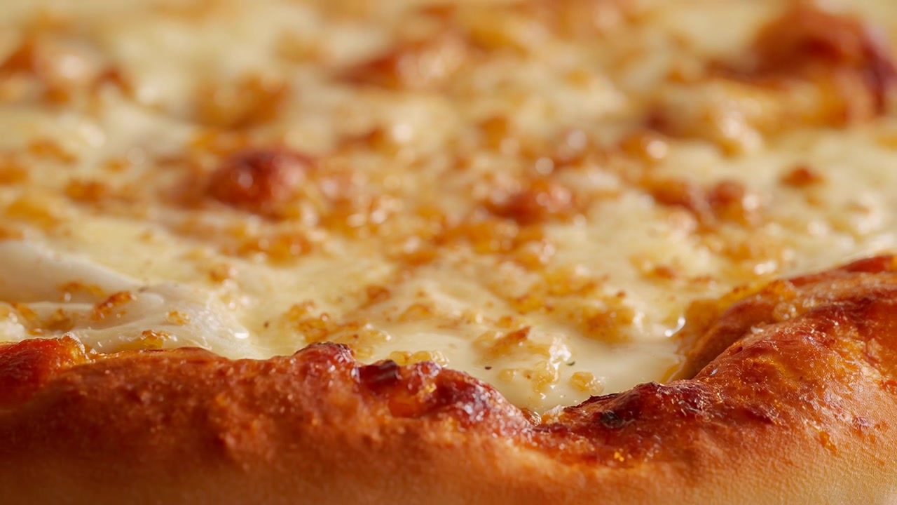 La telecamera si allontana rivelando la consistenza di una pizza al formaggio su un tavolo: formaggio fuso, briciole, olio e crosta dorata.